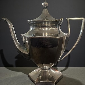 May include: A silver-colored teapot with a geometric design. The teapot has a lid with a finial, a curved spout, and a square handle. The base is also geometric. The teapot is likely made of metal and reflects the dark background.