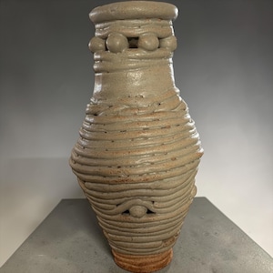 May include: A handmade ceramic vase with a textured, wrapped design. The vase is a light tan color with a reddish-brown base. The top of the vase features a series of spherical accents. The vase has a unique, organic shape.