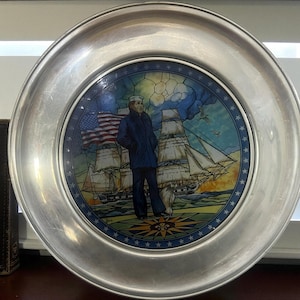 May include: A decorative plate with a silver rim featuring a nautical scene. The center depicts a sailor in a blue coat, an American flag, and sailing ships. The plate has a star-studded border and a compass rose design.