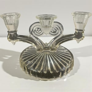 May include: A clear glass candelabra with three candle holders. The base is round with a ribbed design. The arms of the candelabra have a decorative swirl design. The candle holders are cylindrical.
