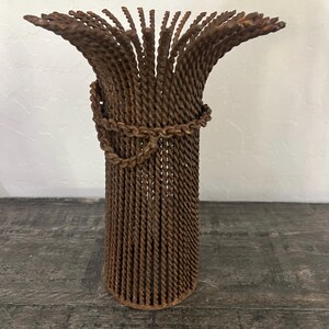 May include: A rustic, brown metal vase with a cylindrical body and flared top. The vase is constructed from twisted metal rods, with a decorative chain detail around the middle. The design gives it a unique, textured appearance.