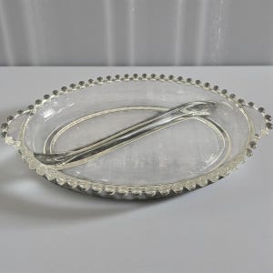 May include: An oval-shaped, clear glass serving dish with a silver-colored serving utensil. The rim of the dish is decorated with small, clear glass beads. The serving utensil is curved and rests inside the dish.