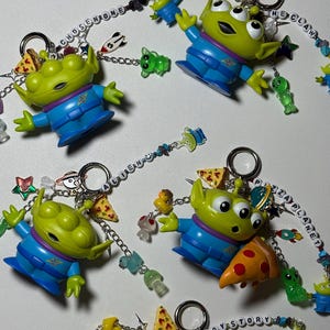 May include: Several green alien keychains with blue outfits and three eyes are displayed. Each keychain has a silver ring and is adorned with charms like pizza slices, small aliens, and the words "Chosen One", "The Claw", "Pizza Planet", and "Toy Story".