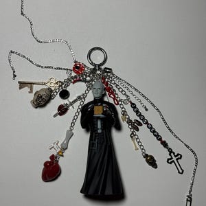 May include: A silver-toned keychain featuring a central Pinhead figure holding a gold cube, surrounded by various charms including a red heart, a skull, a key, a cross, and beads spelling JESUS WEPT, all attached to multiple silver chains.