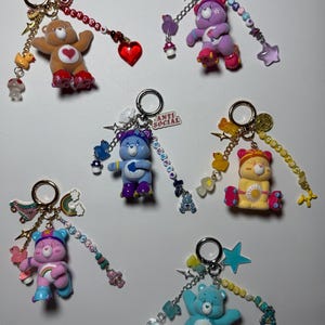 Könnte beinhalten: Sechs Schlüsselanhänger mit Care-Bear-Figuren auf Rollschuhen, jeweils an einem silbernen Metallring befestigt. Die Anhänger bestehen aus verschiedenen Perlen, kleinen Kunststofftieren, Pilzen, Sternen und Buchstabenperlen.