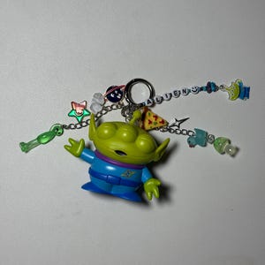 May include: A green alien figurine with a blue and purple outfit, attached to a keychain. The keychain includes charms of a pizza slice, a star, and the word "ALIEN" spelled out with letter beads. Additional charms include a green alien and a blue alien.