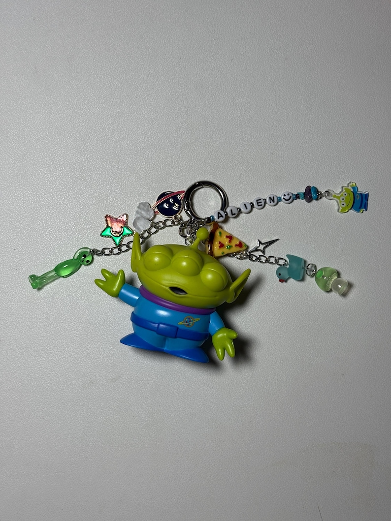 May include: A green alien figurine with a blue and purple outfit, attached to a keychain. The keychain includes charms of a pizza slice, a star, and the word "ALIEN" spelled out with letter beads. Additional charms include a green alien and a blue alien.