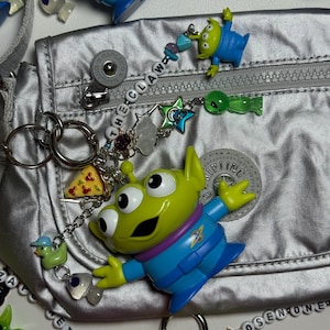 May include: A silver fanny pack with a zipper, surrounded by various charms and keychains. The charms include green alien figures, a pizza slice, and a duck. The text "THE CLAW" is spelled out on a charm. The overall theme is inspired by the movie Toy Story.