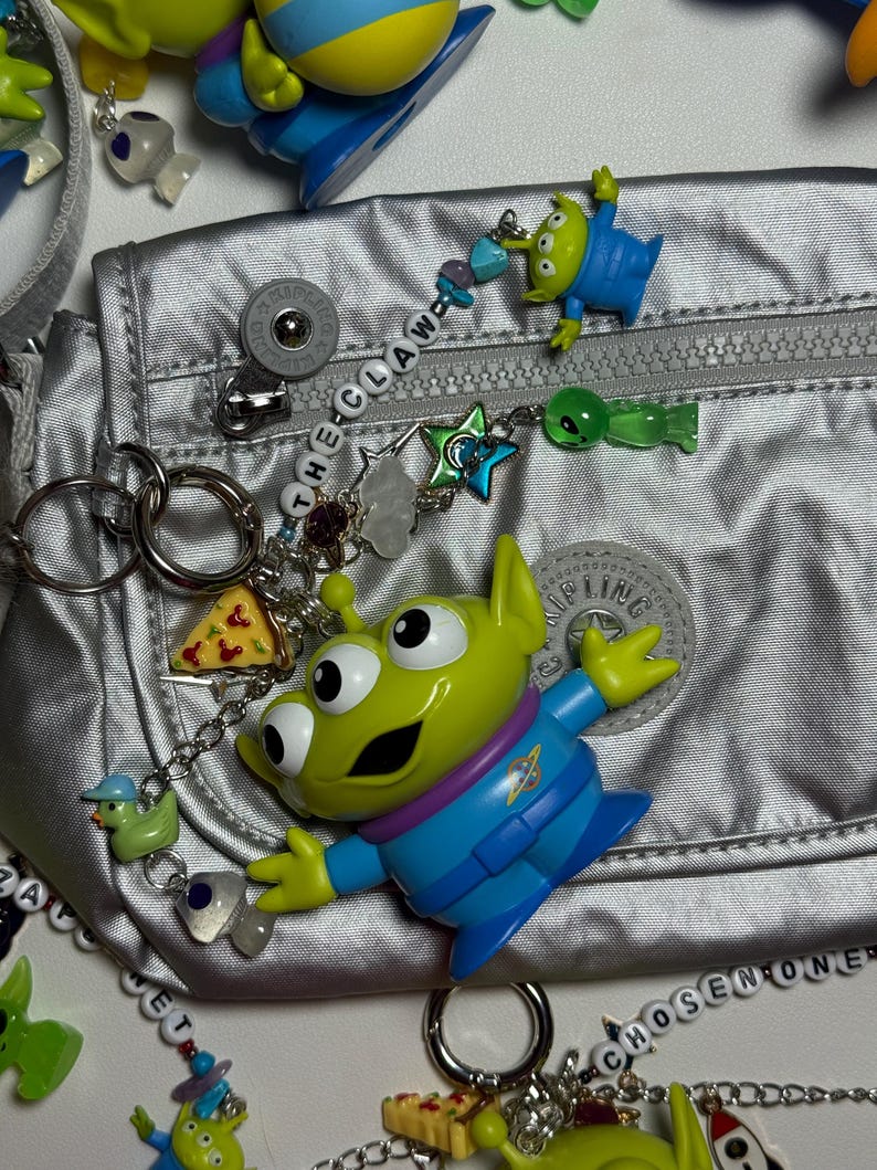 May include: A silver fanny pack with a zipper, surrounded by various charms and keychains. The charms include green alien figures, a pizza slice, and a duck. The text "THE CLAW" is spelled out on a charm. The overall theme is inspired by the movie Toy Story.