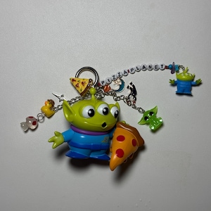 May include: A keychain featuring a green alien character holding a pizza slice. The keychain includes additional charms of a duck, a green alien, and a slice of pizza. The word "PIZZA PLANET" is spelled out in colorful beads.