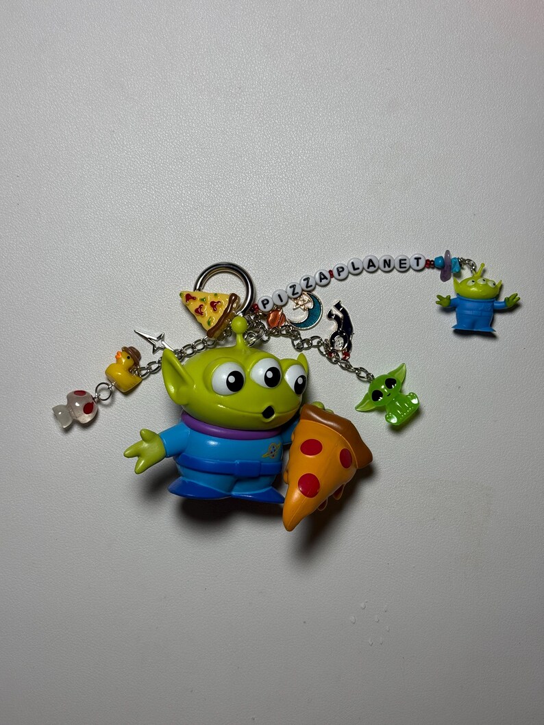 May include: A keychain featuring a green alien character holding a pizza slice. The keychain includes additional charms of a duck, a green alien, and a slice of pizza. The word "PIZZA PLANET" is spelled out in colorful beads.