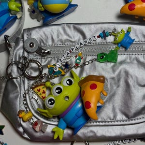 May include: A silver fanny pack with a zipper, surrounded by Toy Story alien charms and keychains. The charms include pizza slices, rockets, and the words "PIZZA PLANET" and "CHOSEN ONE" spelled out with beads. The aliens are green with three eyes.