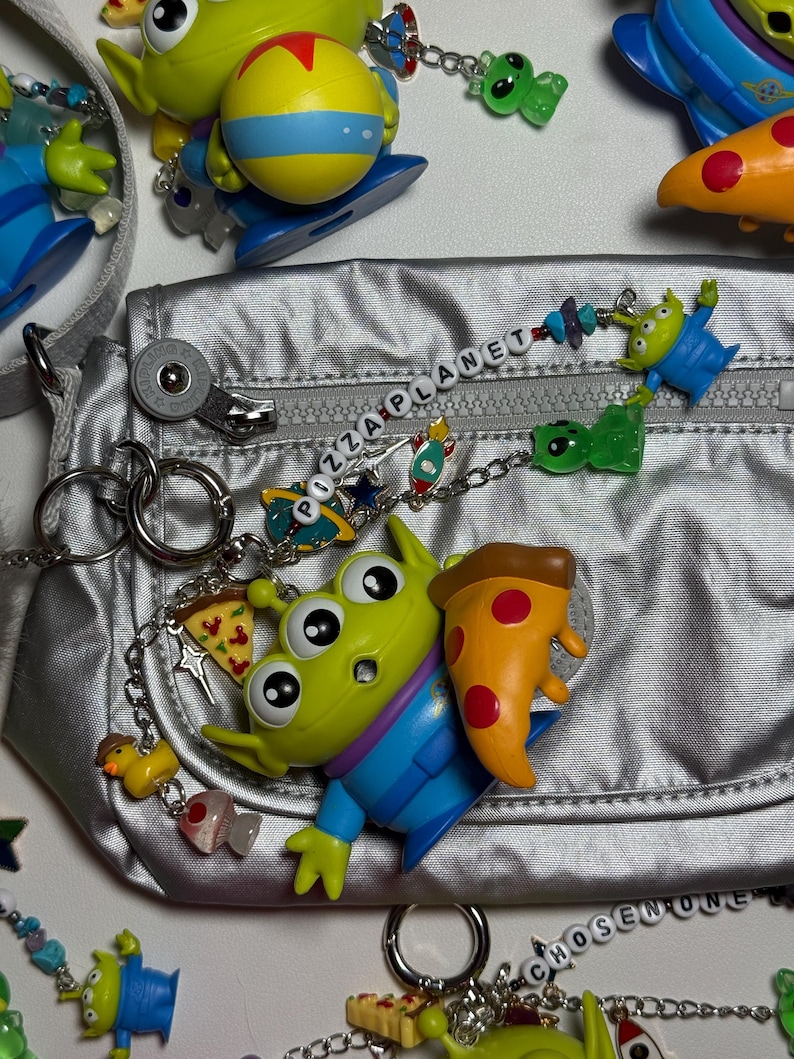 May include: A silver fanny pack with a zipper, surrounded by Toy Story alien charms and keychains. The charms include pizza slices, rockets, and the words "PIZZA PLANET" and "CHOSEN ONE" spelled out with beads. The aliens are green with three eyes.