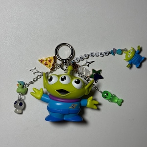May include: A green alien keychain with three eyes, wearing a blue jumpsuit, and the text "THE CLAW". The keychain includes charms of a pizza slice, stars, a duck, a mushroom, and other aliens.
