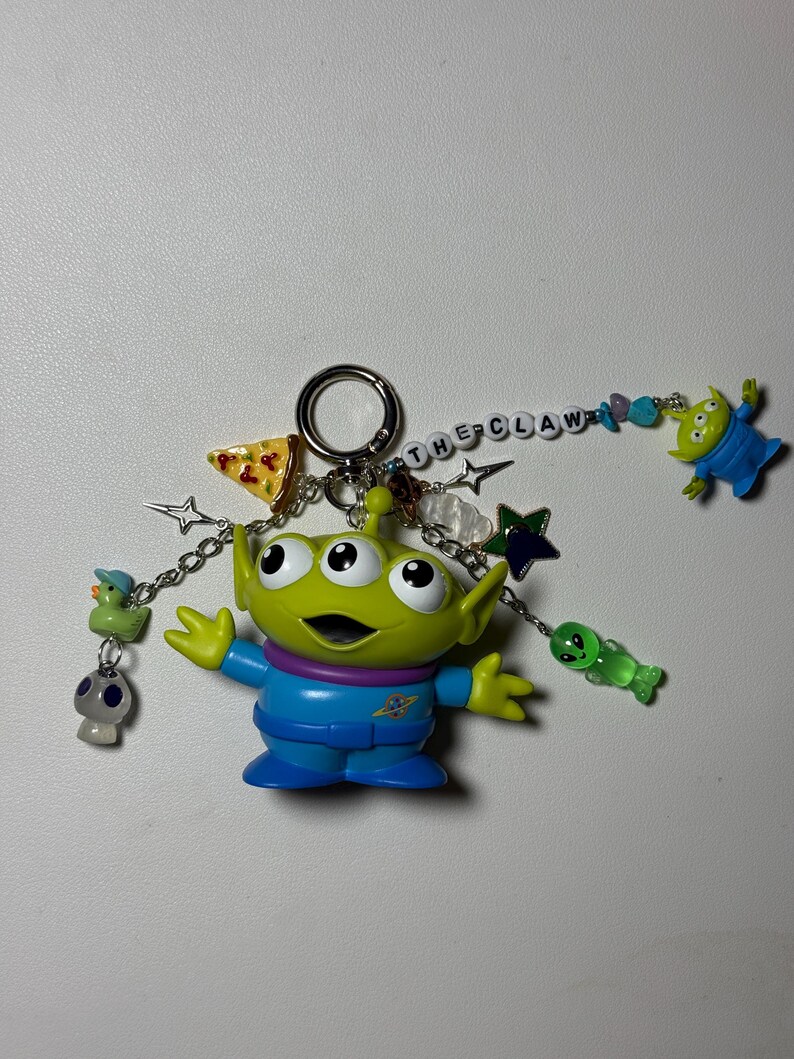 May include: A green alien keychain with three eyes, wearing a blue jumpsuit, and the text "THE CLAW". The keychain includes charms of a pizza slice, stars, a duck, a mushroom, and other aliens.