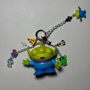 May include: A keychain featuring a green alien figure with blue clothing, surrounded by various charms. The charms include a slice of pizza, stars, and small alien figures. The word "CHOSEN ONE" is spelled out on a chain.