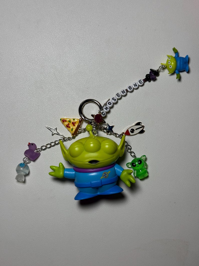 May include: A keychain featuring a green alien figure with blue clothing, surrounded by various charms. The charms include a slice of pizza, stars, and small alien figures. The word "CHOSEN ONE" is spelled out on a chain.