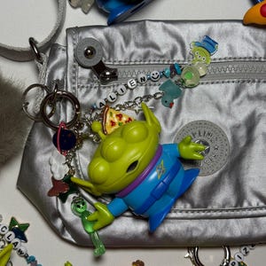 May include: A silver nylon bag with a variety of charms and keychains. The charms include green alien figures, pizza slices, and other colorful designs. The word "ALIEN" is spelled out in beads on a chain.