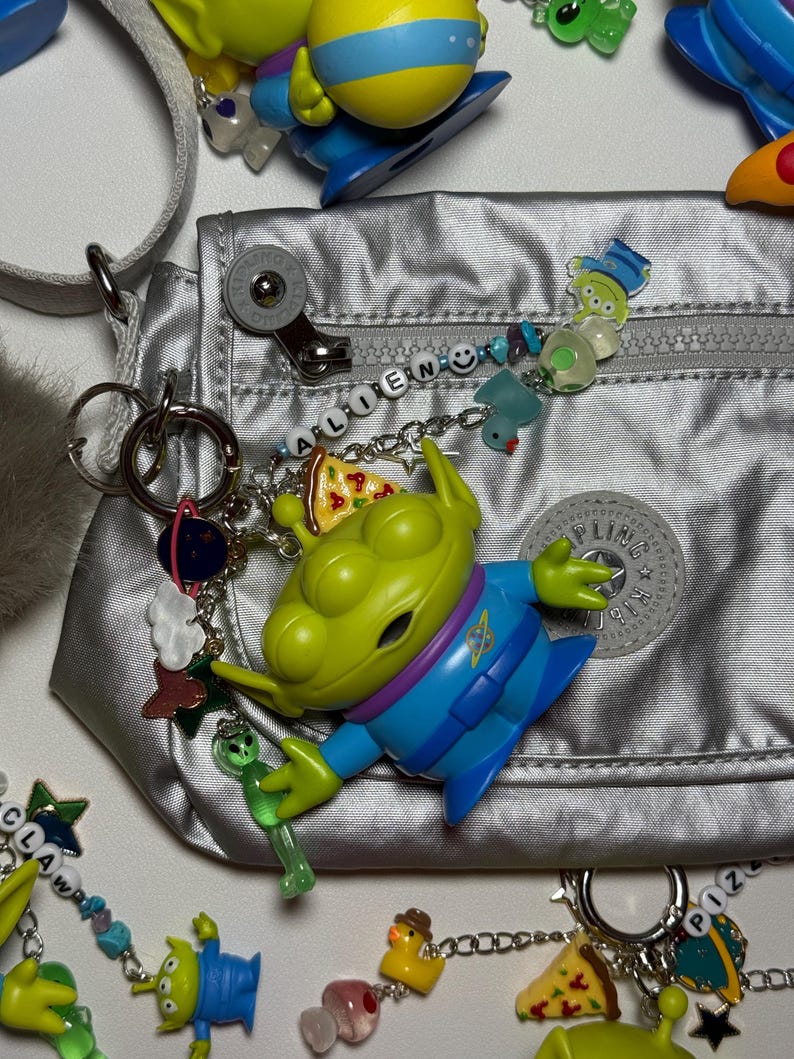 May include: A silver nylon bag with a variety of charms and keychains. The charms include green alien figures, pizza slices, and other colorful designs. The word "ALIEN" is spelled out in beads on a chain.