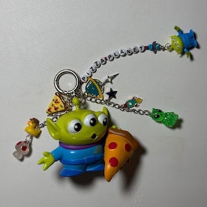 May include: A keychain featuring a green alien holding a pizza slice. The keychain includes various charms, such as a duck, mushroom, and a Pizza Planet banner. Additional alien figures are also attached.