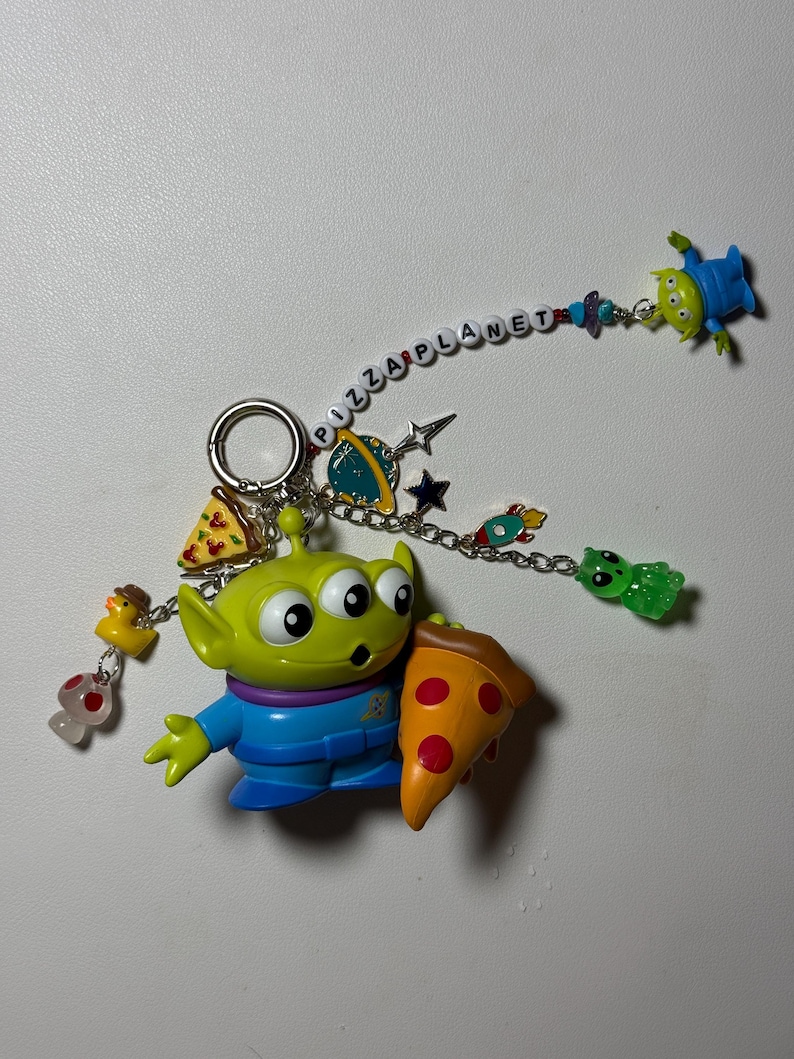 May include: A keychain featuring a green alien holding a pizza slice. The keychain includes various charms, such as a duck, mushroom, and a Pizza Planet banner. Additional alien figures are also attached.