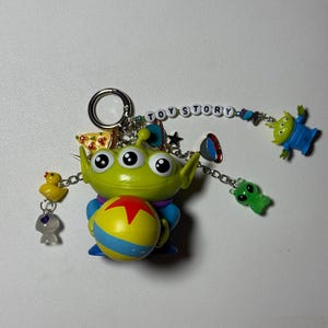 May include: A Toy Story-themed keychain featuring a green alien holding a yellow and blue ball with a red star. The keychain includes additional charms of a yellow duck, a gray alien, and the words "TOY STORY".