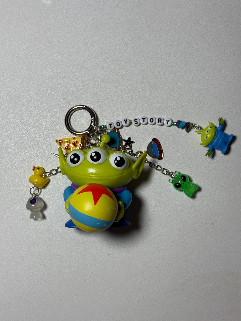 May include: A Toy Story-themed keychain featuring a green alien holding a yellow and blue ball with a red star. The keychain includes additional charms of a yellow duck, a gray alien, and the words "TOY STORY".