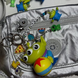 May include: A silver fanny pack is adorned with various Toy Story-themed charms and keychains. The charms include green aliens, a pizza slice, a yellow duck, and a ball with a red star. The word "TOY STORY" is spelled out on a beaded chain.