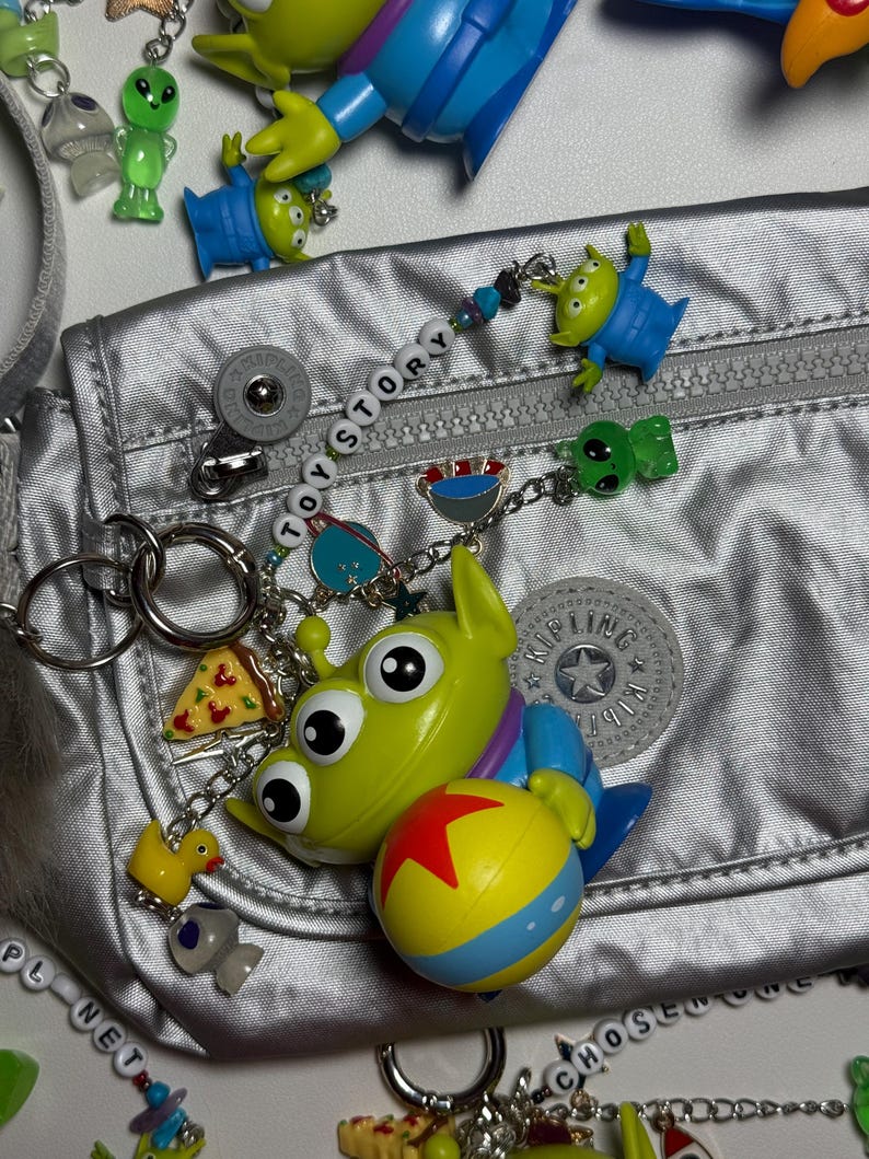 May include: A silver fanny pack is adorned with various Toy Story-themed charms and keychains. The charms include green aliens, a pizza slice, a yellow duck, and a ball with a red star. The word "TOY STORY" is spelled out on a beaded chain.