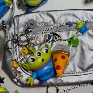 May include: Silver fanny pack with "PIZZA PLANET" beaded zipper pull. Includes Toy Story alien keychains, pizza slice charms, and other themed accessories. The bag has a silver strap and a button closure.