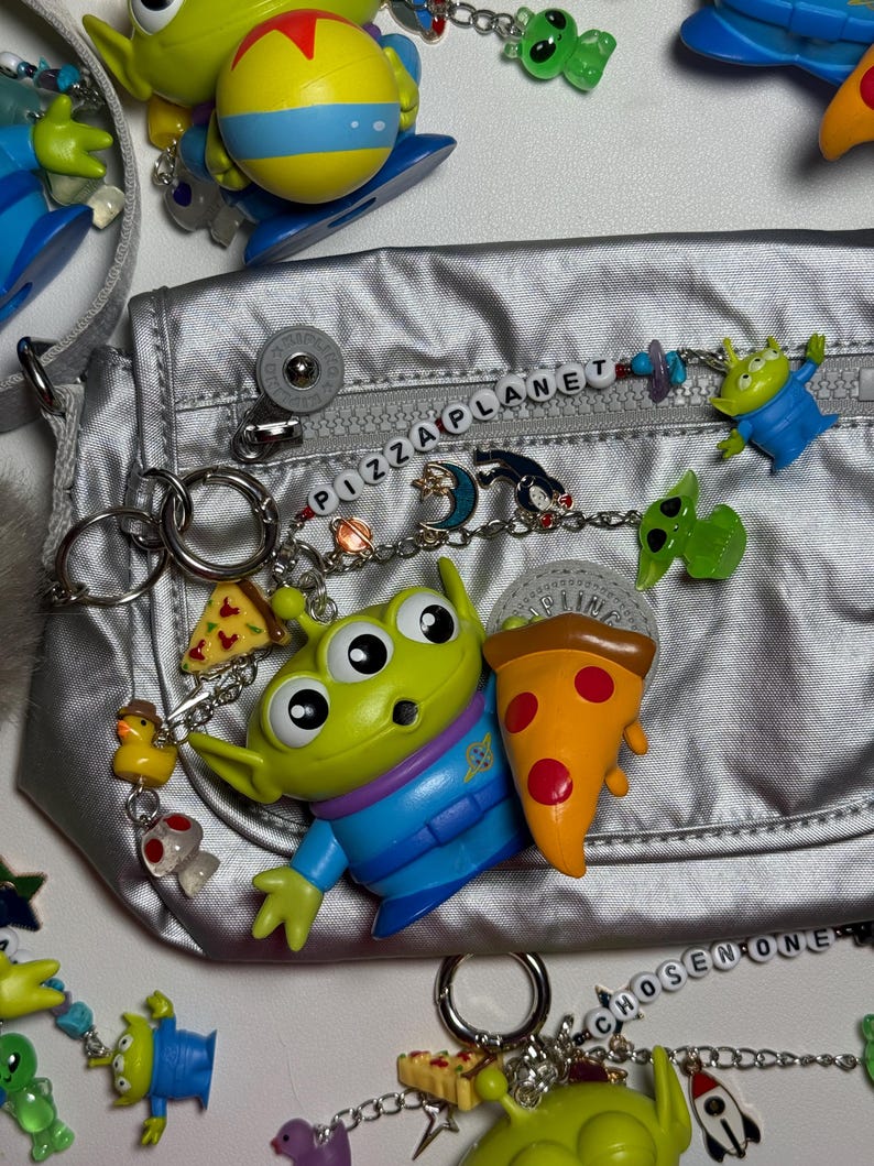 May include: Silver fanny pack with "PIZZA PLANET" beaded zipper pull. Includes Toy Story alien keychains, pizza slice charms, and other themed accessories. The bag has a silver strap and a button closure.