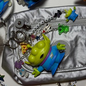 May include: A silver bag is adorned with various charms, including green alien figures, pizza slices, and the text "CHOSEN ONE." The charms are attached to the bag with chains and key rings. The bag has a zipper and a circular logo.