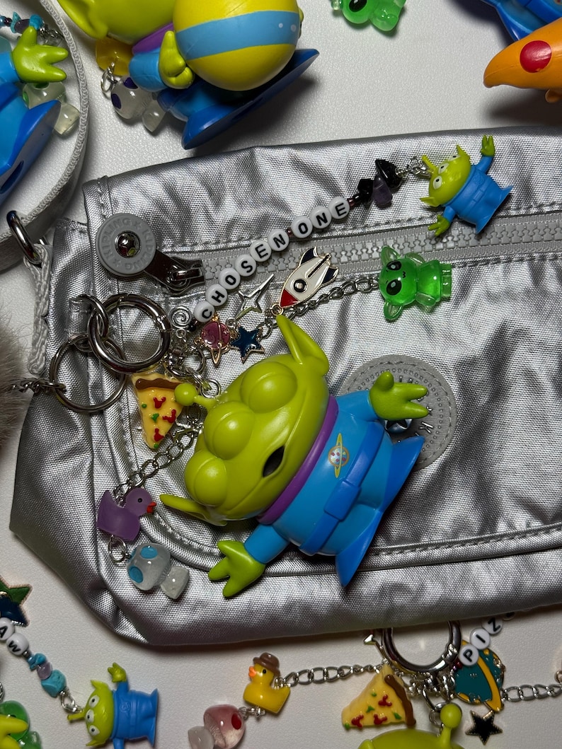 May include: A silver bag is adorned with various charms, including green alien figures, pizza slices, and the text "CHOSEN ONE." The charms are attached to the bag with chains and key rings. The bag has a zipper and a circular logo.
