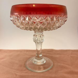 May include: A vintage champagne coupe with a ruby red rim and a clear, diamond-cut bowl. The glass sits on a clear, ornate stem and a circular base. The coupe is likely made of glass and is suitable for serving champagne or other beverages.