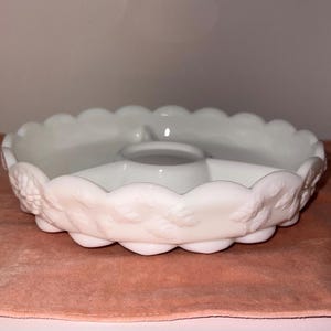 May include: A white, divided serving dish with a scalloped edge and a central dip bowl. The dish features a raised floral design around the rim. The dish is made of opaque glass and sits on a pink surface.