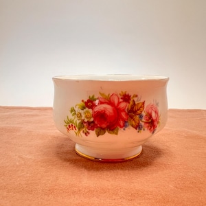 以下が含まれることがあります： 赤いバラ、緑の葉、その他のカラフルな花をあしらった花柄の白いセラミックボウル。 ボウルは金色の縁と台座ベースを備えています。