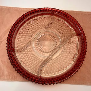 Può includere: Un piatto da portata in vetro trasparente con bordo e divisori rosso rubino. Il piatto è rotondo con un motivo a diamante al centro e un bordo strutturato. Il piatto è progettato per contenere più alimenti.