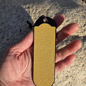 Puede incluir: Un marcapáginas rectangular con una superficie texturizada de color amarillo dorado y una parte superior negra. El marcapáginas tiene un lazo de cuero para colgar. El artículo se sostiene en una mano sobre un fondo neutro.