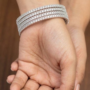 May include: A close-up of a silver bracelet with multiple rows of small, sparkling stones. The bracelet is worn on a wrist, showcasing its intricate design and brilliance. The stones reflect light, creating a dazzling effect.