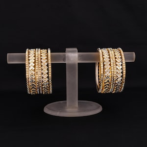 Puede incluir: Dos juegos de brazaletes dorados con patrones geométricos y piedras claras incrustadas se exhiben en un soporte transparente sobre un fondo negro. Los brazaletes están apilados, mostrando sus intrincados diseños y texturas.