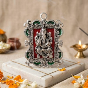 May include: A silver-colored framed religious icon featuring a seated deity with multiple arms, set against a red background. The frame has green and silver accents. The icon is displayed on a white marble surface with gold lines, surrounded by flowers and a lit candle.