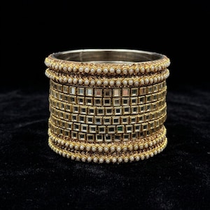 K&ouml;nnte beinhalten: Goldfarbenes Armreif-Armband mit mehreren Schichten. Das Armband hat kleine quadratische Spiegelakzente und ist von Reihen kleiner, wei&szlig;er, perlenartiger Perlen umrandet. Der Hintergrund ist schwarz.