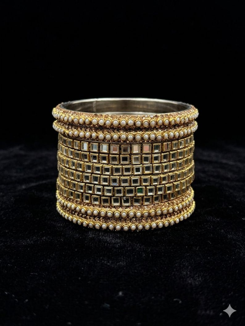 K&ouml;nnte beinhalten: Goldfarbenes Armreif-Armband mit mehreren Schichten. Das Armband hat kleine quadratische Spiegelakzente und ist von Reihen kleiner, wei&szlig;er, perlenartiger Perlen umrandet. Der Hintergrund ist schwarz.