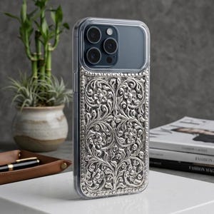 May include: A protective phone case for an iPhone 17 Pro featuring a metallic silver back panel with an intricate, raised floral filigree pattern. The case has a clear border and a cutout for the camera lenses. Text reads: The Silver Filigree Collection - for iPhone 17 Pro.