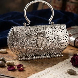May include: Ornate silver-colored handbag with a curved handle. The bag features intricate floral and leaf patterns, with a decorative clasp and small dangling bells along the bottom edge. The bag is on a wooden surface.