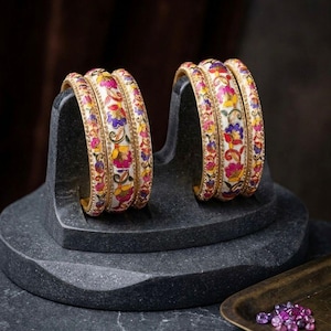 Può includere: Un set di sei bracciali rigidi con base bianca e complessi motivi floreali nei toni del rosa, viola, giallo e verde. Ogni bracciale presenta un bordo color oro metallizzato e un profilo liscio e arrotondato, disposti in due gruppi.