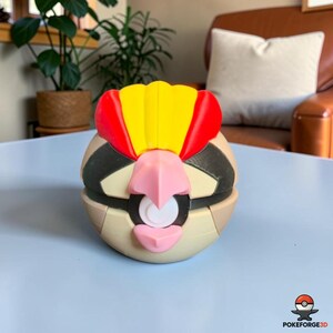 May include: A spherical, multi-colored plastic figure resembling a bird-like creature with a yellow and red crest, a pink beak, and a central white circular button on a black band. The object features tan, black, and cream-colored panels.