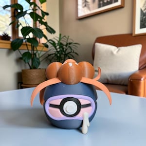 May include: A 3D-printed, stylized figure in shades of blue, pink, and brown, resembling a cartoon character. It features a large, circular eye, curved orange appendages, and a white, teardrop-shaped detail. The figure is set on a light blue surface.
