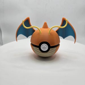 May include: An orange and blue Charizard-themed Poké Ball. The ball features a round design with a white button, brown accents, and orange wings with blue tips. The wings are attached to the top of the ball with yellow lines.