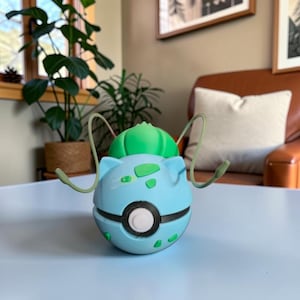 May include: A light blue and green Bulbasaur-themed Poké Ball. The ball has a black stripe around the middle and a white button. Green accents and two green tendrils extend from the top. The product is on a white surface.